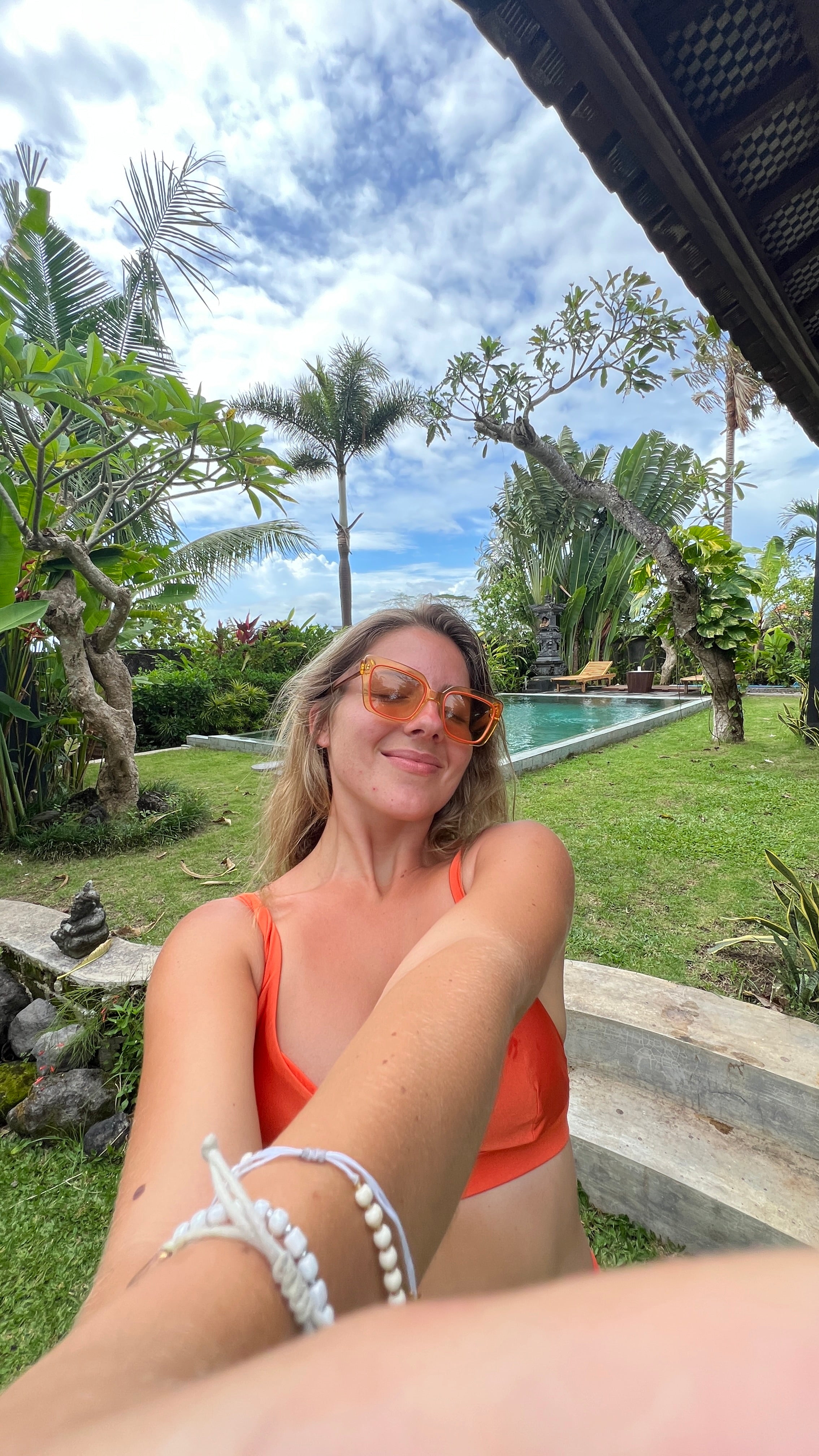 Smiling woman in sunny garden wearing orange sunglasses, enjoying a tropical getaway with lush greenery.